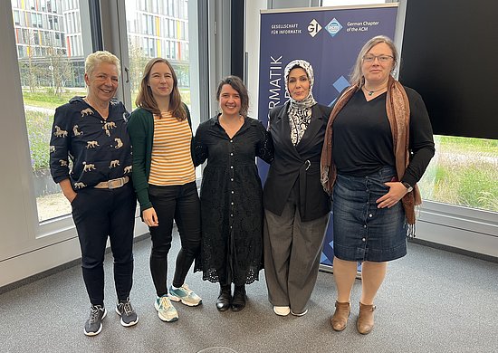Fünf frauen stehen nebeneinander und haben einander im Arm. Sie stehen vor einer Fensterfront und einem Aufsteller der GI.