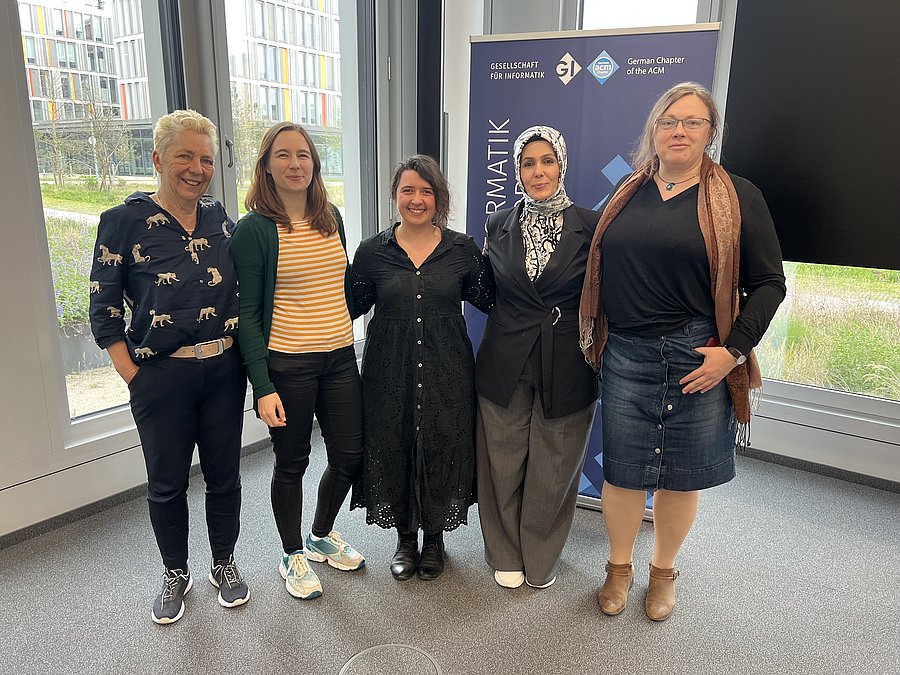 Fünf frauen stehen nebeneinander und haben einander im Arm. Sie stehen vor einer Fensterfront und einem Aufsteller der GI.
