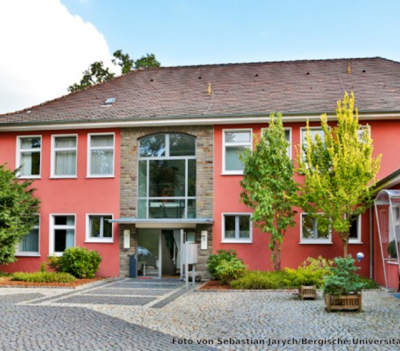 Rot gestrichenes Gebäude mit Schrägdach und grünen Bäumen davor. Der Platz vor dem Eingang ist mit Kopfsteinen geflastert. Der Himmel im Hintergrund ist blau.
