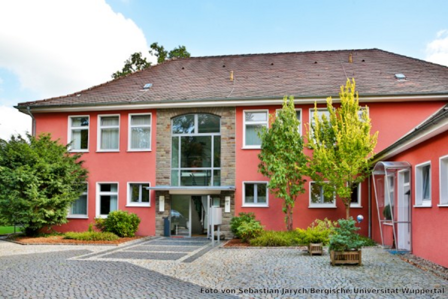 Rot gestrichenes Gebäude mit Schrägdach und grünen Bäumen davor. Der Platz vor dem Eingang ist mit Kopfsteinen geflastert. Der Himmel im Hintergrund ist blau.