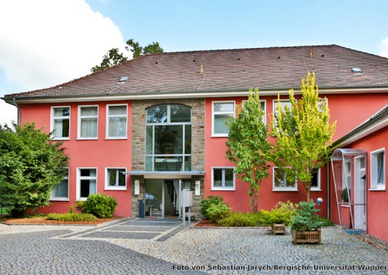 Rot gestrichenes Gebäude mit Schrägdach und grünen Bäumen davor. Der Platz vor dem Eingang ist mit Kopfsteinen geflastert. Der Himmel im Hintergrund ist blau.