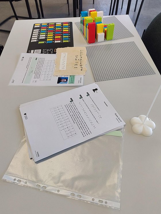 Ein weißer Stationstisch der Science Quest mit Braille Legosteinen in verschiedenen Farben, Legoplatten, einer Braille Übersetzungstafel sowie mehrere Arbeitsblättern und einem Lösungswort in Braille auf einem brauen Zettel.