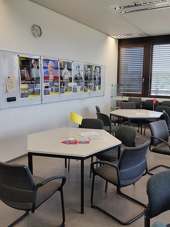 Ein Besprechungsraum mit einzelnen Gruppentischen, Fenstern im Hintergrund und einem Whiteboard auf der linken Seite. Auf den Tischen stehen Fähnchen in den Farben Grün, Gelb und Rot sowie Arbeitsblätter und diverse andere Arbeitsmaterialien. Am Whiteboard hängen sechs verschiedene Poster berühmter Informatiker*innen sowie ein Kreuzwerträtsel auf braunem Papier.