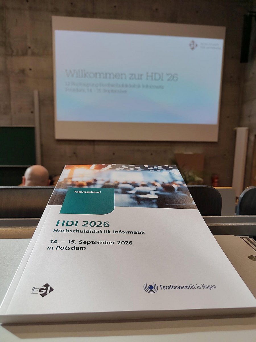 Der Tagungsband der HDI liegt auf einem Tisch im Hörsaal. Im Hintergrund ist eine Willkommens-Folie zu sehen.