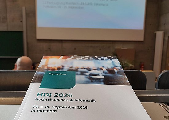 Der Tagungsband der HDI liegt auf einem Tisch im Hörsaal. Im Hintergrund ist eine Willkommens-Folie zu sehen.